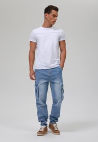 Weißes T-Shirt mit kurzen Ärmeln und hellblauen Cargo-Hosen mit seitlichen Taschen, kombiniert mit braunen High-Top-Sneakers. Glatte Textur und lässige Passform.