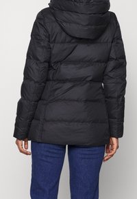 Musta toppatakki hupulla, jossa tikattu kuvio, joustavat hihansuut ja hieman kaareva helma. Yhdistetty sinisiin farkkuihin.