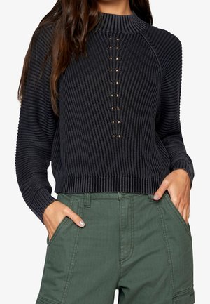 Femme portant un pull en maille côtelée noir avec des détails de boutons à l'avant et un pantalon vert taille haute, les mains dans les poches.