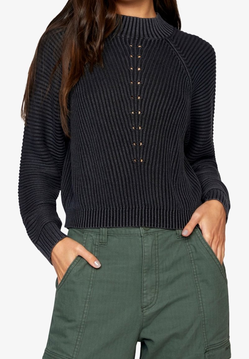 Femme portant un pull en maille côtelée noir avec des détails de boutons à l'avant et un pantalon vert taille haute, les mains dans les poches.