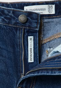 Jean en denim bleu foncé avec une braguette ouverte, bouton marqué "Stradivarius", étiquette de taille 38, et étiquette intérieure portant l'inscription "SOIS TA PROPRE MUSE."