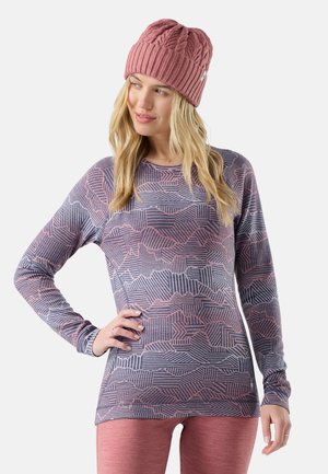 Mujer con gorro de punto rosa, camiseta de manga larga con estampado en azul y rosa, y mallas rosas, de pie con una mano en la cadera.