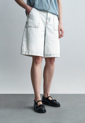 Osoba oblečená v svetlých rifľových krátkych nohaviciach po kolená s veľkými prednými vreckami, modrý top a čierne topánky typu Mary Jane, stojaca na sivom podlaží.