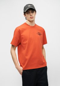 Νεαρός άνδρας που φοράει πορτοκαλί μπλουζάκι Dickies και σκούφο Dickies σκουρόγριου χρώματος, στέκεται με το ένα χέρι στην τσέπη μπροστά σε απλό φόντο.