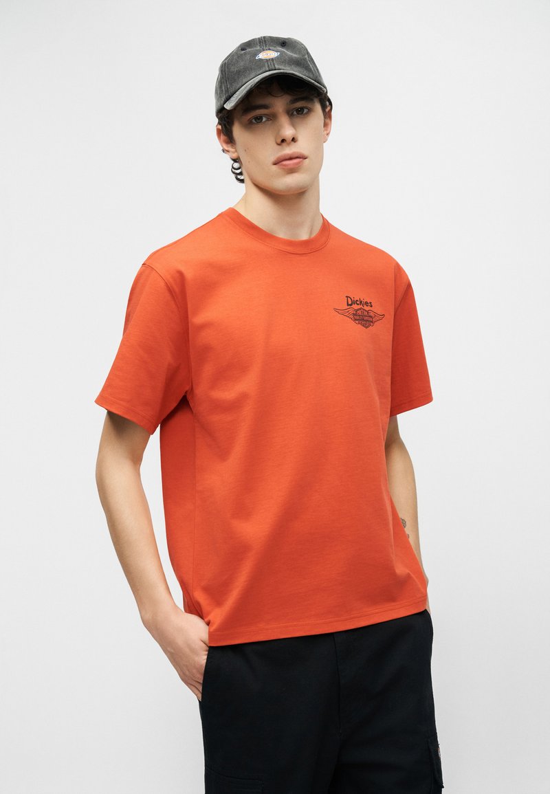 Junger Mann, der ein oranges Dickies-T-Shirt und eine dunkelgraue Dickies-Kappe trägt, steht mit einer Hand in der Tasche vor einem einfarbigen Hintergrund.