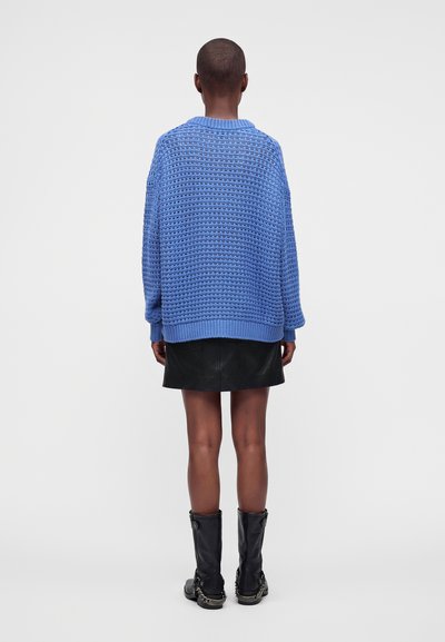 Personne aux cheveux courts portant un pull texturé bleu, une jupe en cuir noire et des bottes noires mi-mollet, debout de dos sur un fond uni.