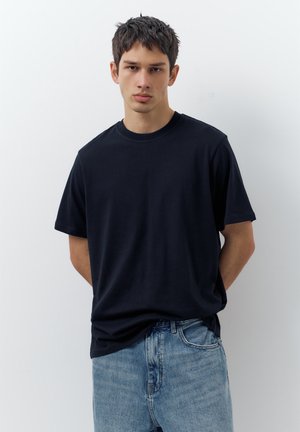 Young man with short dark hair wearing a plain black t-shirt and light blue jeans standing against a white background.