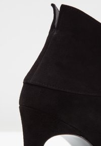Bottine en daim noir avec un talon bloc incurvé et une languette à l'arrière, présentée sur un fond blanc.