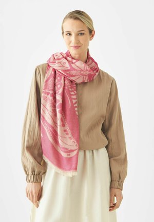 Femme portant un chemisier beige et une jupe blanche, avec une grande écharpe rose à motifs enveloppée autour du cou et tombant sur son épaule.