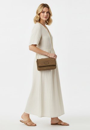 Vrouw in een beige jurk tot aan de enkels en sandalen staat zijwaarts, met een bruine crossbodytas met kettingband over haar schouder.