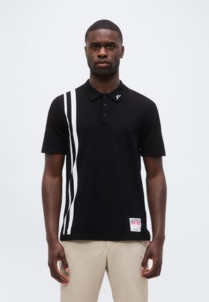 Mand iført sort polo-shirt med hvide lodrette striber på venstre side og beige bukser, stående mod en ensfarvet lys baggrund.