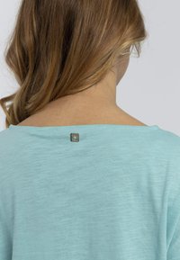 Top azzurro chiaro a maniche corte con scollo ampio. Presenta un piccolo dettaglio del logo argentato sul retro del collo. Texture morbida e liscia.