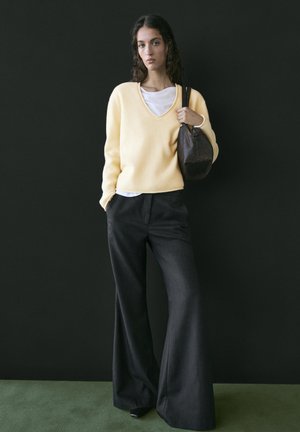 Kvinde iført en sart gul V-hals trøje, hvid skjorte under, sorte bukser med brede ben og smalle striber, bærende en mørkebrun lædertaske over den ene skulder.