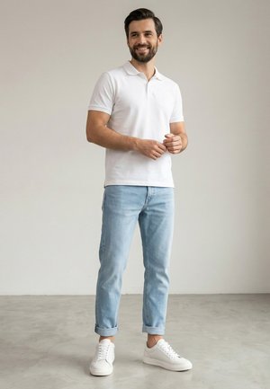 Mężczyzna z brodą, ubraną w białe polo, jasnoniebieskie podwinięte jeansy i białe trampki, stojący na szarej podłodze na tle jednolitej ściany.