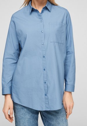 Persoon draagt een lichtblauw overhemd met lange mouwen, knopen aan de voorkant en een borstzak, gecombineerd met een blauwe spijkerbroek, tegen een effen achtergrond.