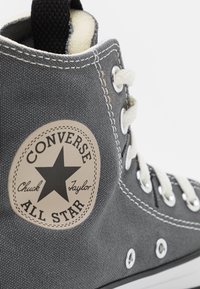 Šedá plátěná vysoká teniska s bílou gumovou podrážkou, černou štítkovou tabulkou, kulatou nášivkou s černou hvězdou a textem "Converse All Star".