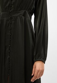 Σκούρο πράσινο σατέν φόρεμα με κουμπωτό μπροστινό �μέρος, συγκεντρωμένη μέση και μακρύ μανίκι με ελαστικές μανσέτες. Λείο υλικό, κομψός σχεδιασμός.