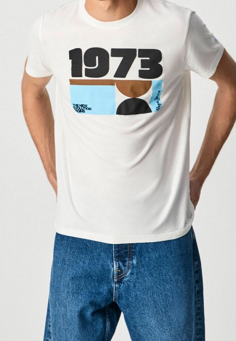 Mand iført en hvid T-shirt med "1973" og abstrakte farveblokke, parret med blå denim jeans, hænderne hvilende på hoften.