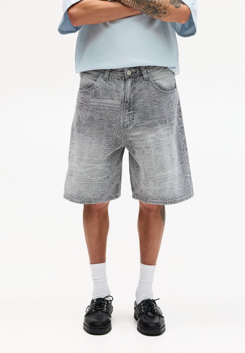 Shorts di denim grigi con motivi profondi, vestibilità rilassata, design a cinque tasche e chiusura con bottone. Abbinati a scarpe nere e calzini bianchi.