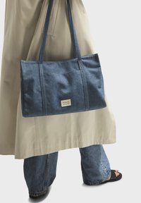 Personne portant un trench-coat beige et un jean bleu, tenant un sac cabas en denim bleu avec une étiquette "nat & nin", debout sur un fond blanc.