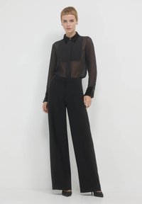 Schwarze, transparente Bluse mit Knöpfen und Kragen, kombiniert mit weit geschnittenen schwarzen Hosen mit seitlichen Akzenten und hohen Schuhen.
