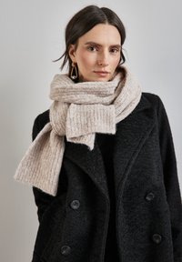 Beige striklet tørklæde med ribbet tekstur, viklet om halsen. Parret med en sort, dobbeltknappet frakke med store knapper.
