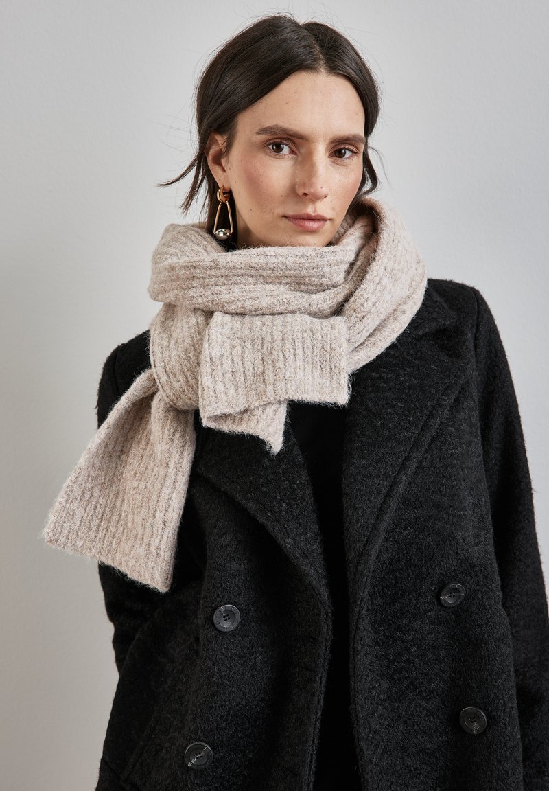 Beige striklet tørklæde med ribbet tekstur, viklet om halsen. Parret med en sort, dobbeltknappet frakke med store knapper.
