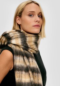 Écharpe en tissu à carreaux noirs, beiges et crème, fabriquée en laine douce. Présente un design surdimensionné et confortable avec une texture duveteuse.