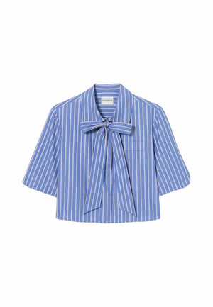Mujer con blusa azul a rayas con lazo, falda negra de cuero, calcetines beige, zapatos marrones, sosteniendo un bolso de ante marrón sobre un fondo liso.