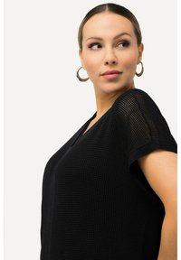 Černý top s krátkým rukávem vyrobený z texturované síťoviny s výstřihem do V. Má volný střih a jemné detaily šití.