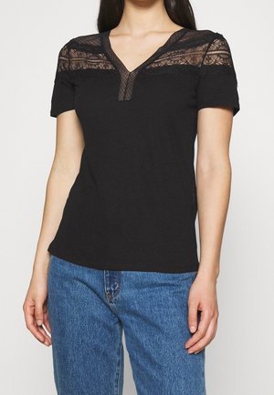 Mujer vistiendo una blusa negra de manga corta con yugo de encaje y escote en V, combinada con jeans azules, de pie frente a un fondo liso.