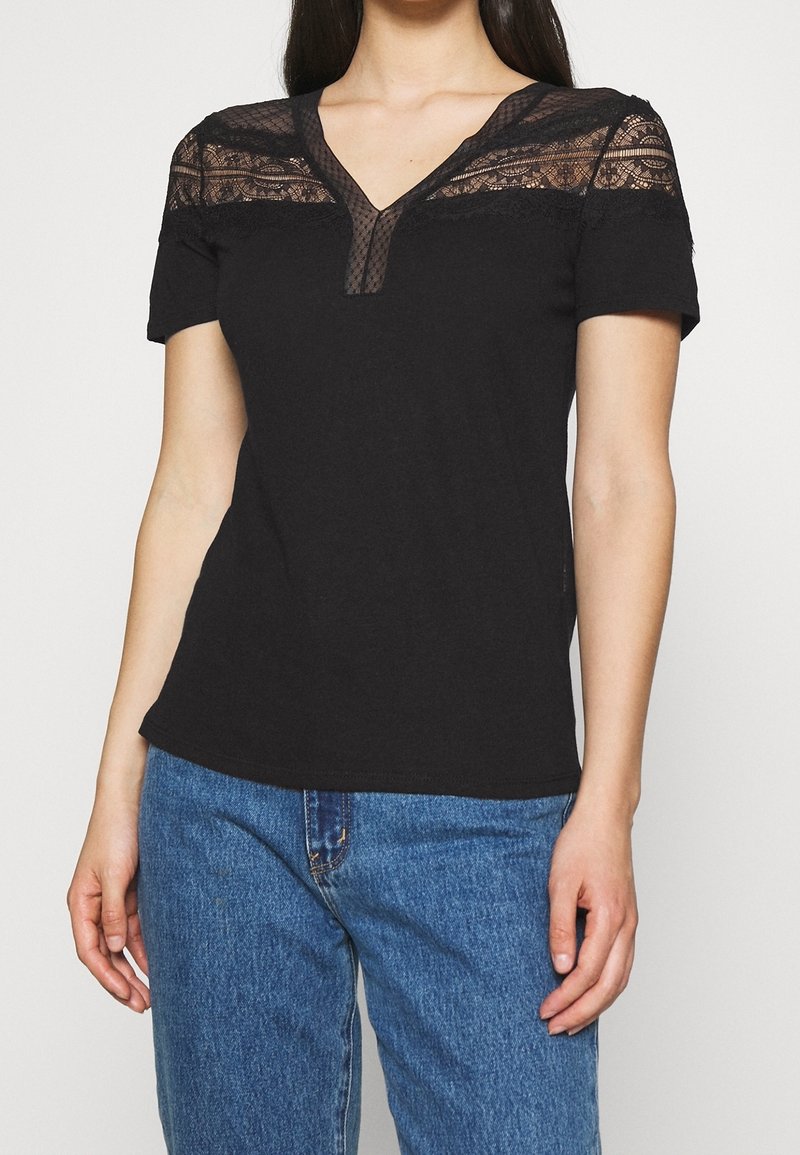 Femme portant un haut noir à manches courtes avec empiècement en dentelle et décolleté en V, associé à un jean bleu, se tenant devant un fond uni.