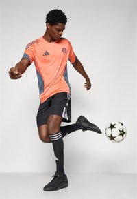 Felrood en grijs sportshirt, zwarte shorts, zwarte schoenen en kniehoge zwarte sokken met witte strepen. Voetbal in de buurt.