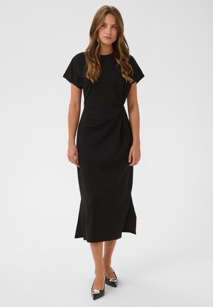 Femme aux cheveux longs portant une robe midi noire à manches courtes et des talons aiguilles noirs, debout devant un fond uni.