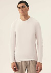 Weißes Langarmhemd aus glattem Stoff, runder Ausschnitt und taillierte Passform. Getragen mit hellgrauen Jogginghosen.