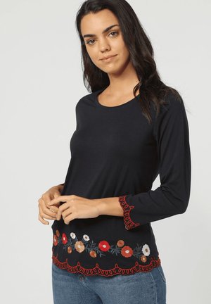 Camisa negra de manga larga con bordado floral en rojo, blanco y marrón en el dobladillo. Acabado texturizado en las mangas y borde inferior ondulado.
