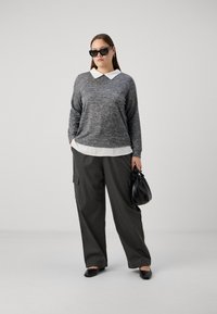 Suéter gris de punto con cuello blanco, superpuesto a una camisa blanca. Pantalones holgados verde oscuro y bolso negro. La modelo lleva gafas de sol negras.