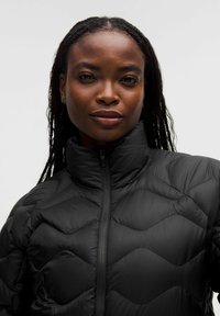 Primer plano de una mujer con el pelo trenzado que lleva una chaqueta acolchada negra con un cuello alto y cremallera frontal, contra un fondo liso.