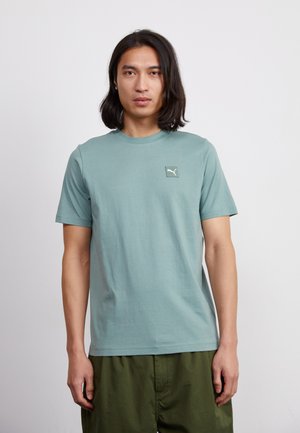 T-shirt in cotone turchese chiaro con colletto girocollo e un piccolo logo Puma ricamato sul lato sinistro del petto, abbinata a pantaloni corti verde oliva.