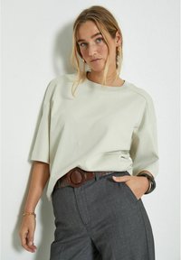 Light gray, oversized cotton shirt with dropped shoulders, round neckline, and short sleeves; paired with dark gray trousers and a brown textured belt.