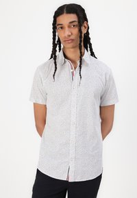 Camicia bianca a maniche corte con un motivo a pois neri. Presenta una patta a righe a contrasto e un orlo curvo.