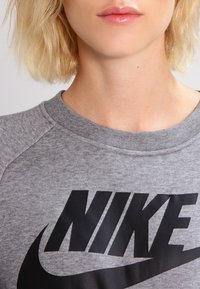 Šedá bavlněná mikina s černým logem Nike přes přední stranu. Má kruhový výstřih a raglánové rukávy. Hladká textura.