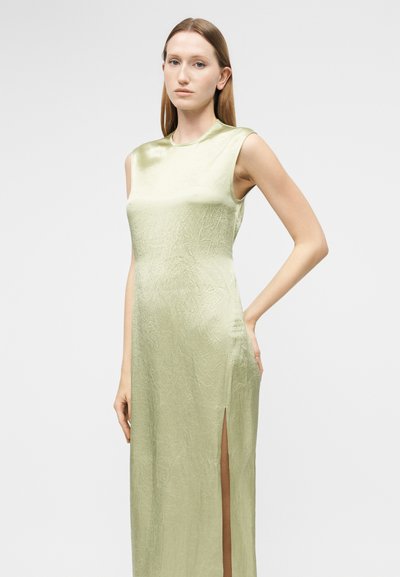 Γυναίκα που φοράει αμάνικο, ανοιχτό πράσινο σατέν φόρεμα με ψηλή πλαϊνή σκίσιμο, στέκεται με το ένα χέρι στη μέση της μπροστά σε απλό φόντο.