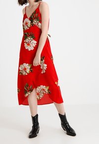 Röd blommig midiklänning med tunna band, prydd med stora vita pioner och gröna blad, kompletterad med en sidoklyvning och ankelboots.