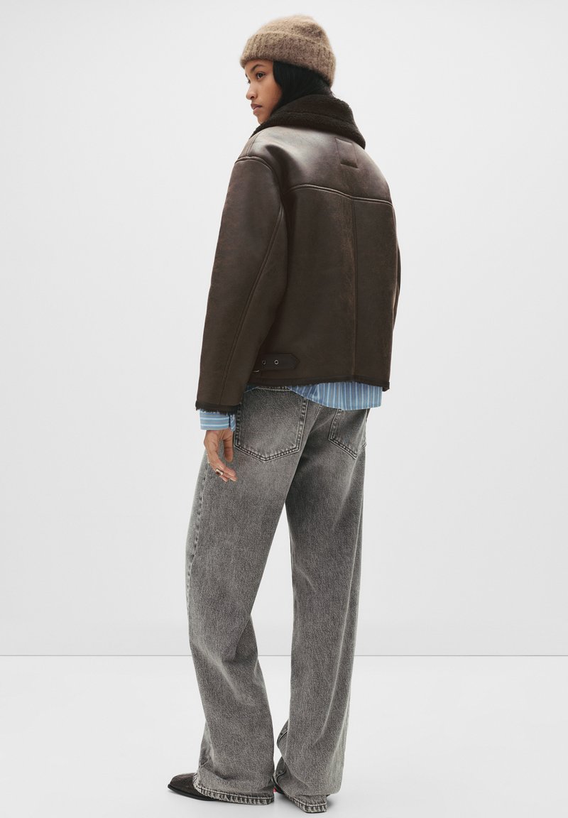 Žena v hnědém kabátu s umělým beránkem, béžové pletené čepici, modro-bílé pruhované košili, volných šedých džínách a černých botách, stojící na bílém pozadí.