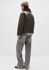 Vrouw draagt een bruine shearling jas, beige gebreide muts, blauw gestreept overhemd, losse grijze spijkerbroek en zwarte schoenen, staand tegen een witte achtergrond.