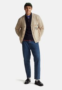 Béžový svetr na knoflíky s copánkovým vzorem, přes něj tmavě modrý svetr na zip, doplněný modrými džínami a černými mokasínami.