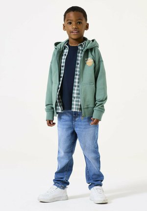 Felpa verde con zip sopra una maglietta blu navy, camicia a quadri e jeans in denim azzurro chiaro. Le sneakers bianche completano l'outfit.