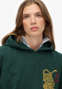 Jonge vrouw met lichtbruin haar, gekleed in een donkergroene hoodie met een gele geborduurde tijger en rode Chinese tekens.