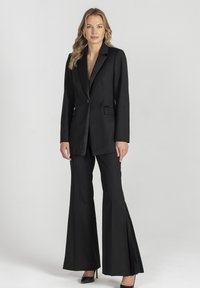 Czarny, dopasowany garnitur z długą marynarką zapinaną na jeden guzik oraz dopasowanymi rozkloszowanymi spodniami z plisowanymi detalami. Całość uzupełniają krótkie buty na obcasie.